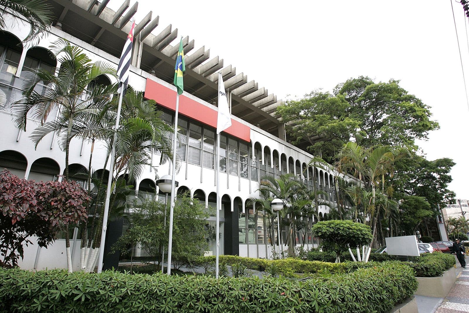 Fachada do prédio da Imprensa Oficial do Estado de São Paulo, com arcos brancos, bandeiras e palmeiras no jardim da frente