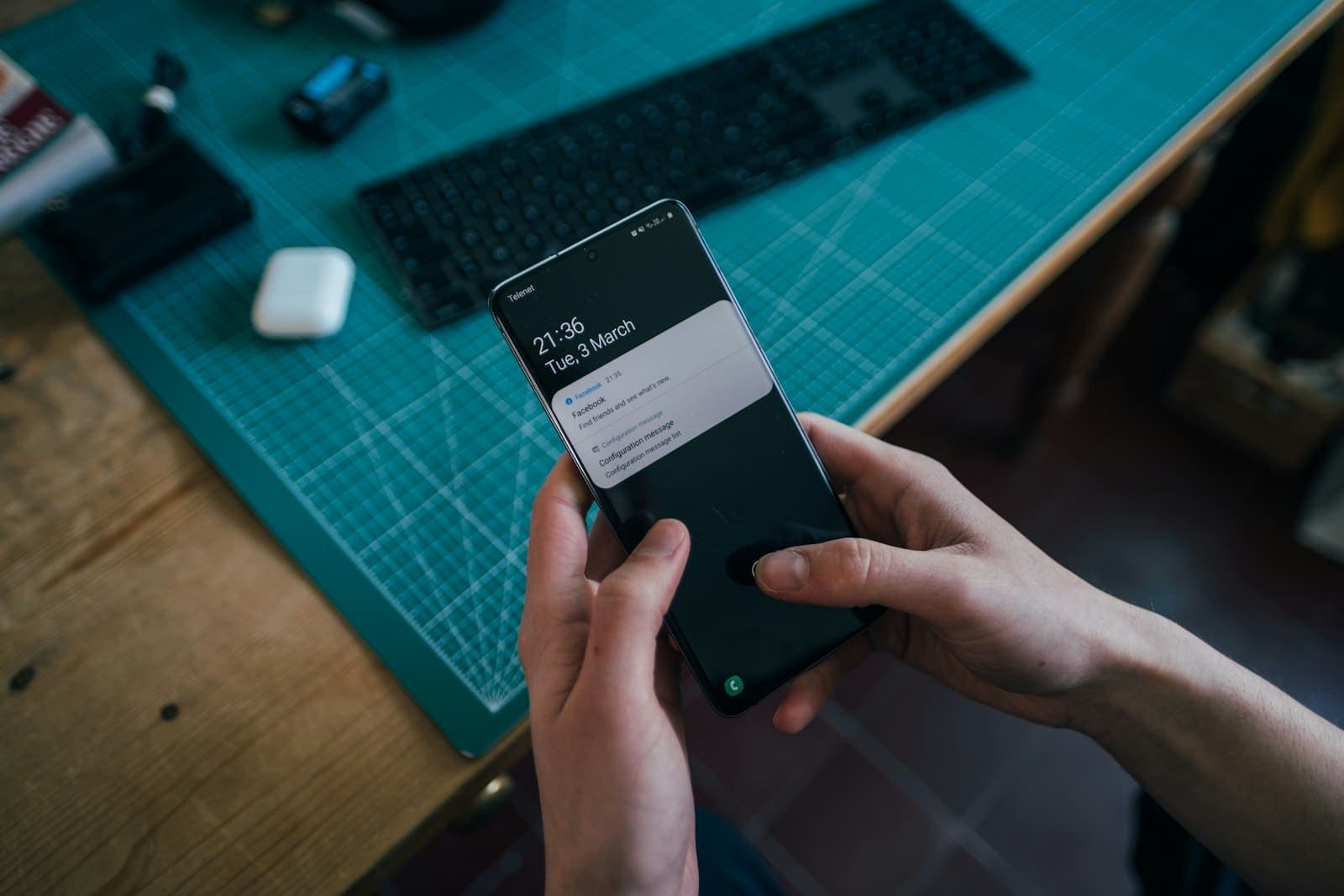 Mãos segurando um celular com notificações na tela, sobre uma mesa de trabalho